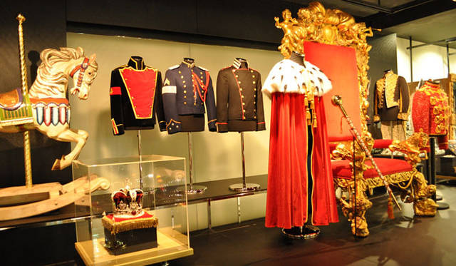 <b>MICHAEL JACKSON - The Official NEVERLAND Collection｜マイケル・ジャクソン ― オフィシャル ネバーランドコレクション</b><br><b>右・ミリタリージャケット</b>　このナポレオンスタイルのロングミリタリーコートは、手の込んだゴールドの糸による装飾が目を引く。アラン・ベネットによるデザイン。<b>中・コスチューム・ケープ</b>　赤いベルベッドでつくられたフルレングスのケープは、着脱可能なにせのオコジョの襟とゴールドのトリミングがほどこされている。また、宝石に似た原石のついたゴールドのメタル製十字架のブローチがついている。デザインは「Dennis Tompkins Michael Bush」によるもの。© MICHAEL JACKSON ESTATE
