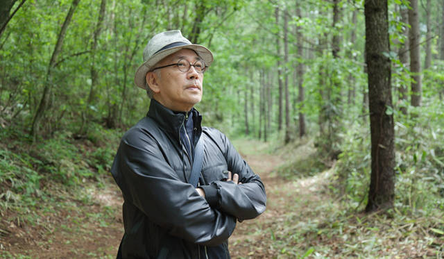 Ryuichi Sakamoto stands amidst the "LOUIS VUITTON FOREST by more trees," now brighter after thinning. "This is my second visit, and the thinned areas are truly luminous. It's visually beautiful, isn't it? Grass is growing below, giving an overall impression of lush greenery."